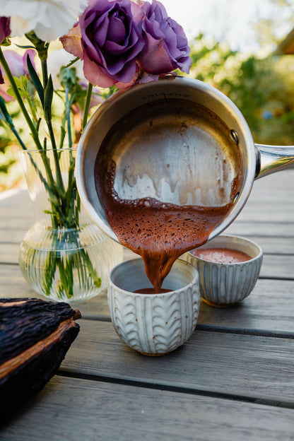 Hot chocolate being poured from a saucepan into a cup on a wooden table with flowers in the background.